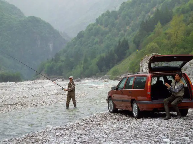 Twee individuen vissen in een ondiepe, rotsachtige rivier met bergen en bomen op de achtergrond; de ene persoon staat in de rivier, terwijl de ander in de open kofferbak van een geparkeerde rode Volvo 850 stationwagen zit.