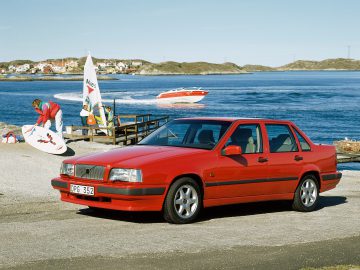 Een rode Volvo 850 sedan staat geparkeerd aan de waterkant met op de achtergrond een windsurfer die uitrusting klaarmaakt en een rode boot die te hard over het water vaart.