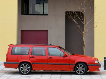 Een rode Volvo 850 stationwagen staat geparkeerd op een verhard terrein voor een modern gebouw met beige, bruine en rode delen. Vlakbij staat een bladerloze boom.