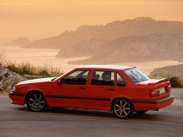 Een rode Volvo 850 sedan staat geparkeerd op een bergweg met uitzicht op een schilderachtig uitzicht op de oceaan bij zonsondergang.