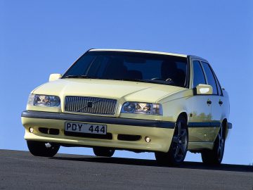 Een gele Volvo 850 sedan met kenteken PDY 444 staat geparkeerd op een hellend oppervlak tegen een helderblauwe lucht op de achtergrond.