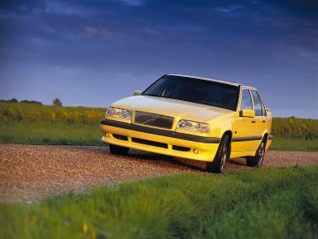 Een Volvo 850 sedan rijdt op een onverharde weg omgeven door groene velden onder een bewolkte hemel.