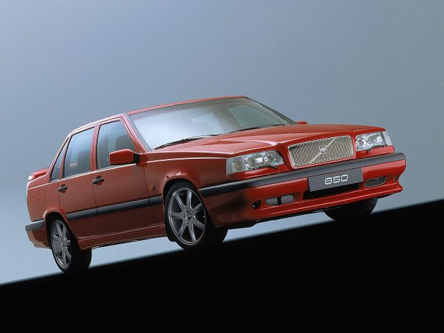 Een rode Volvo 850 sedan is iets schuin geplaatst, waardoor de opvallende grille en het strakke zijprofiel goed tot hun recht komen. De auto valt op tegen een donkere voorgrond met een contrasterende lichtblauwe achtergrond.