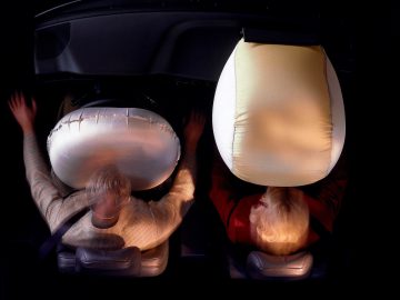 Een bovenaanzicht van twee mensen die in een Volvo 850 zitten met geactiveerde airbags ervoor. Beide personen dragen veiligheidsgordels en de airbags zijn volledig opgeblazen.