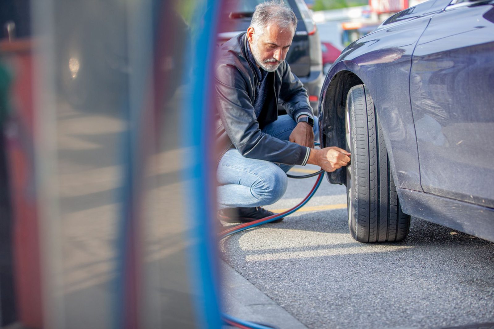 Een man knielt naast een auto en pompt de band op met een luchtpomp bij een benzinestation. Banden oppompen? Zo doe je dat!