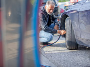 Een man knielt naast een auto en pompt de band op met een luchtpomp bij een benzinestation. Banden oppompen? Zo doe je dat!
