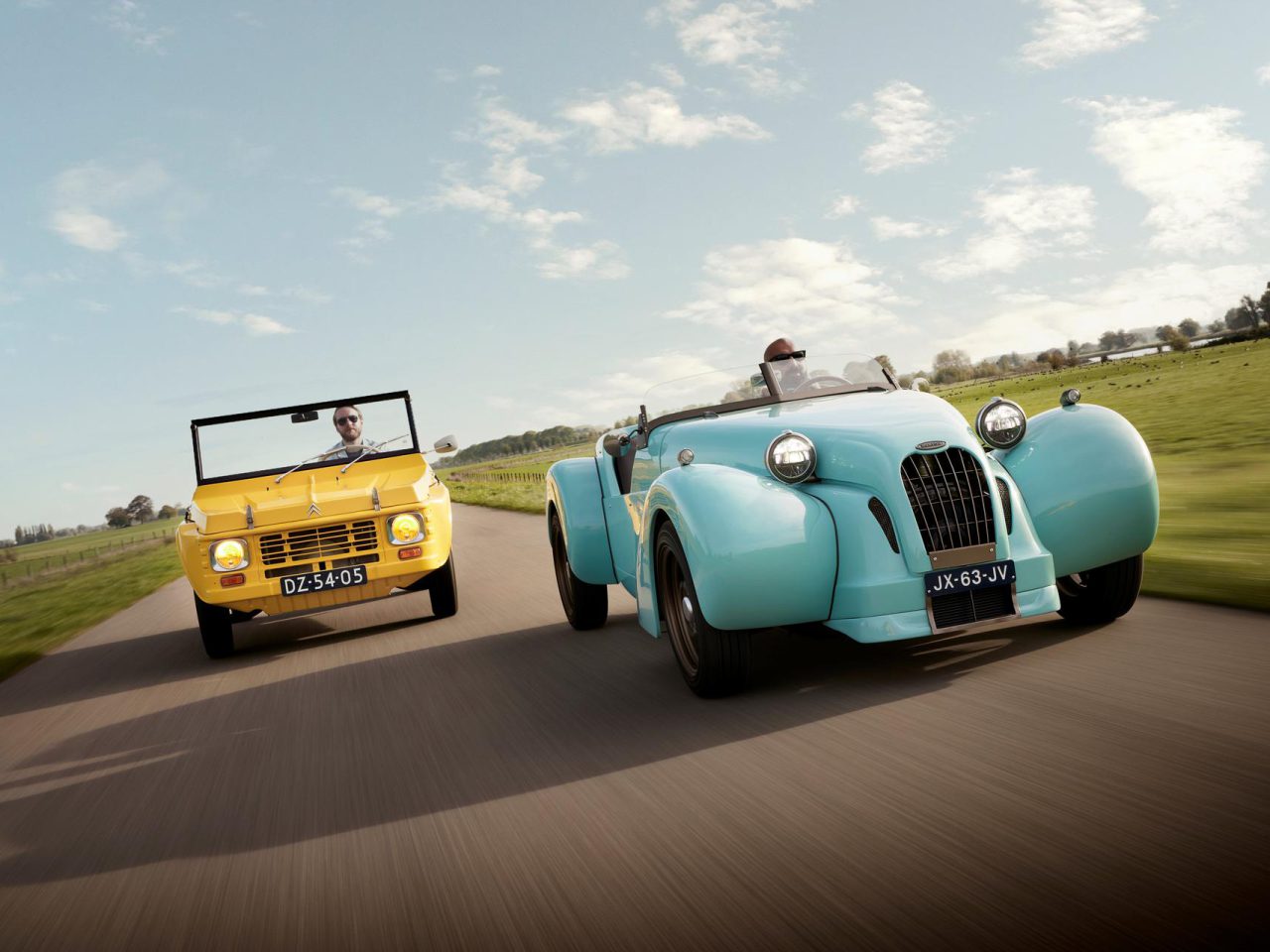 Twee oldtimers, een gele cabriolet en een lichtblauwe cabrioletcoupé, die doet denken aan de klassieke Burton-stijl, rijden op een landelijke weg met aan weerszijden grasvelden onder een gedeeltelijk bewolkte hemel.