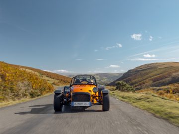 Een Caterham Seven 340 sportwagen rijdt op een smalle plattelandsweg met met gras begroeide heuvels en een helderblauwe lucht op de achtergrond.