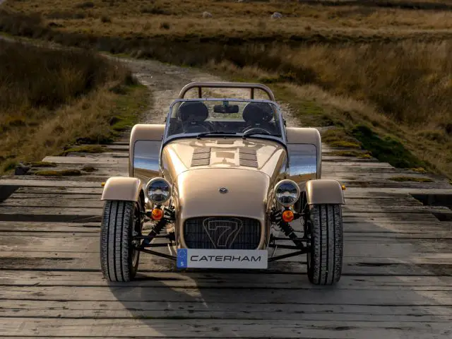 Een lichtbruine Caterham Seven 340 sportwagen staat geparkeerd op een houten brug in een landelijk, grasachtig landschap.