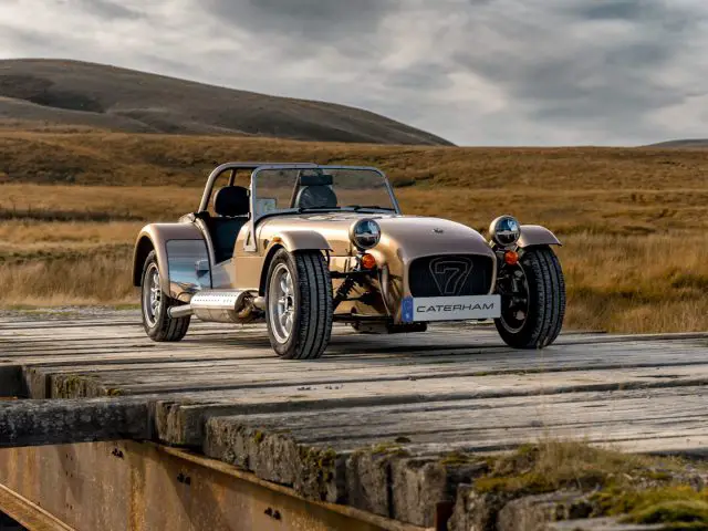 Een Caterham Seven 340-sportwagen staat geparkeerd op een houten brug in een schilderachtig landelijk gebied met glooiende heuvels op de achtergrond.