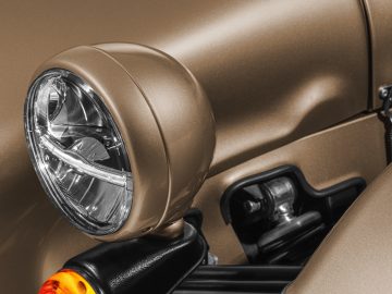 Close-up van een ronde koplamp op een bronskleurige Caterham Seven 340, met een deel van het spatbord en de motorkap van de auto zichtbaar.