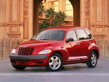 Een rode Chrysler PT Cruiser staat geparkeerd voor een gebouw met palmbomen en stenen muren, dat een unieke mix van retrocharme en moderne sfeer belichaamt.