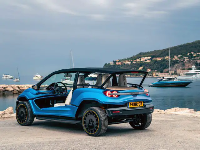 Een blauwe Strandbuggy met open dak staat geparkeerd vlakbij een waterkant met jachten en heuvels op de achtergrond onder een bewolkte hemel.