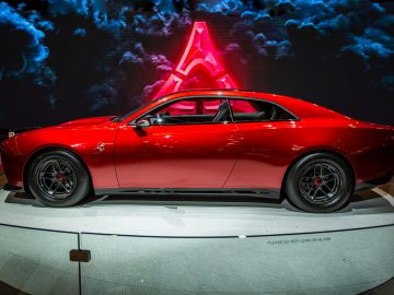 Een slanke, rode tweedeurs Dodge Charger Daytona SRT coupé tentoongesteld tegen een achtergrond met gestileerde A- en wolkenafbeeldingen. Op het bord aan de voorkant staat: "LEUN NIET OP GLAS.