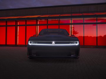 Een slanke, zwarte Dodge Charger Daytona SRT met een verlichte grille staat in de schemering geparkeerd voor een gebouw met rood getinte ramen.