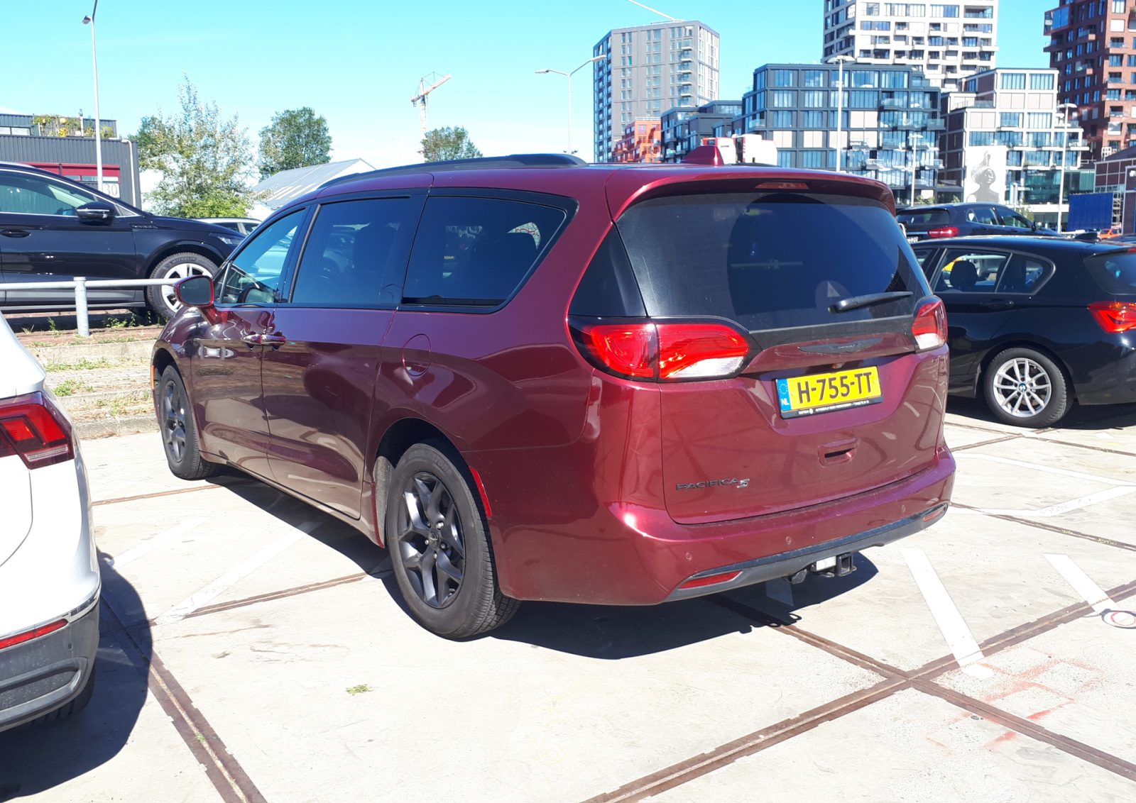 Een rode Chrysler Pacifica-minibus staat geparkeerd naast andere voertuigen met gebouwen op de achtergrond. Op het achterste kenteken staat "H-755-TT".