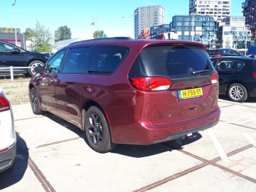 Een rode Chrysler Pacifica-minibus staat geparkeerd naast andere voertuigen met gebouwen op de achtergrond. Op het achterste kenteken staat "H-755-TT".
