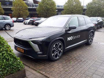 Op een regenachtige dag staat een zwarte SUV met de tekst 'NIO Service' op de zijkant geparkeerd. Laadkabels zijn aangesloten op het voertuig. Op de achtergrond zijn verschillende andere auto's en bomen zichtbaar, wat de inzet van NIO voor duurzame mobiliteit, zelfs bij slecht weer, benadrukt.