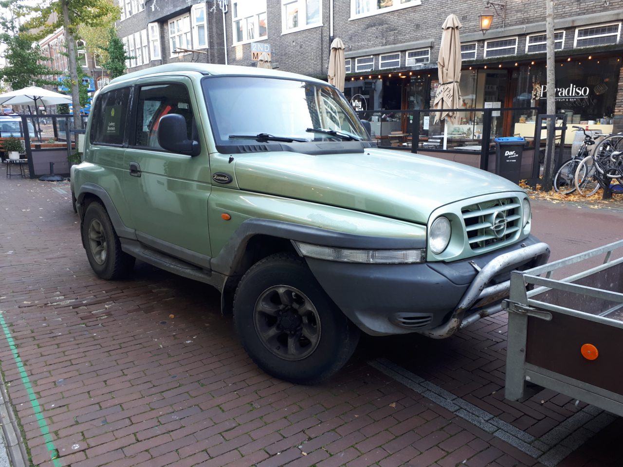 Een lichtgroene Ssangyong Korando met een zwarte bumper en motorkap staat geparkeerd in een geplaveide straat voor een restaurant en enkele fietsen.