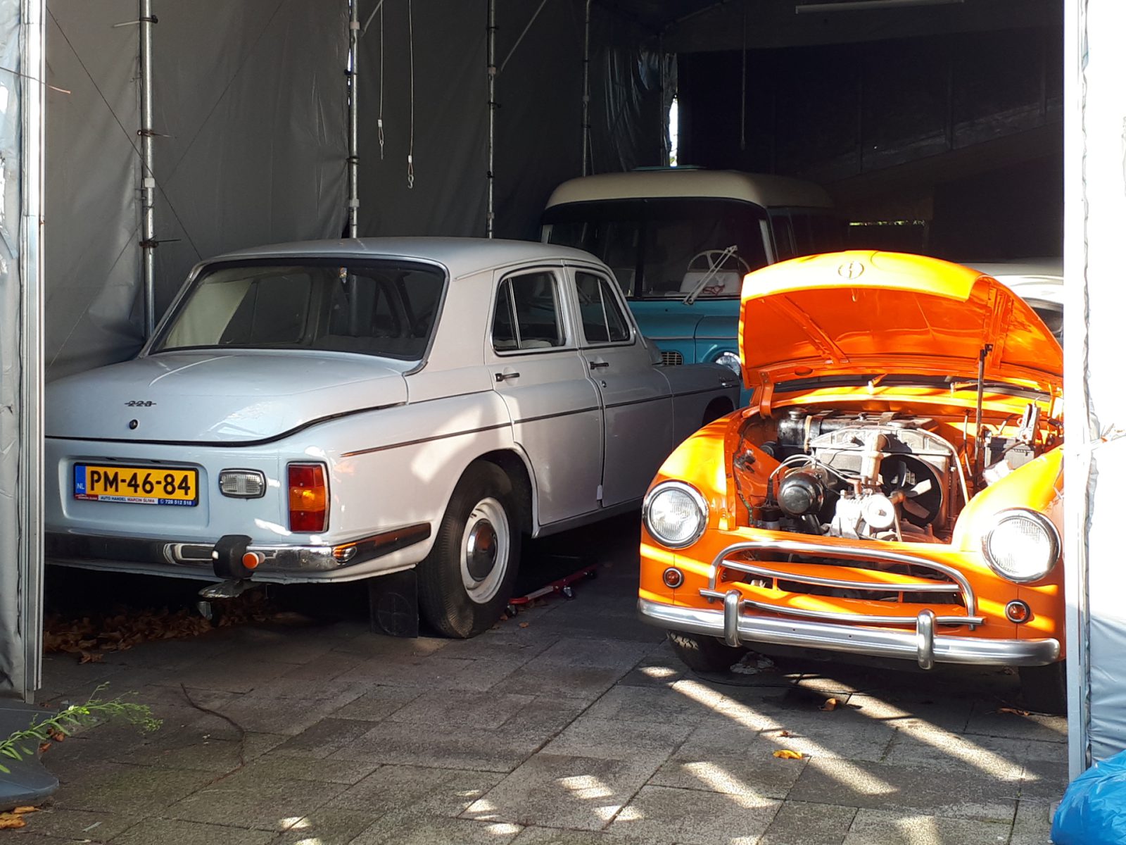 Een oranje klassieke auto met open motorkap, mogelijk een FSO-model, staat naast een witte klassieke auto onder een tent geparkeerd.