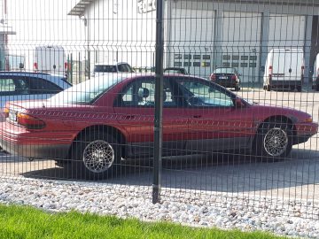 Een rode sedan met zichtbare schade aan de voorbumper staat naast andere voertuigen geparkeerd, achter een hekwerk van gaas. Dit voertuig werd onlangs gespot door verschillende voorbijgangers.