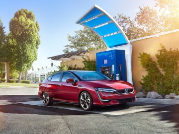 Bij een waterstoftankstation staat een rode sedan geparkeerd met een blauwe overkapping erboven, aangevuld met waterstofpanelen, omringd door bomen en een gebouw op de achtergrond.