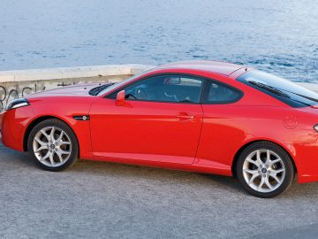 Een vrouw in een zwarte jurk loopt langs een rode Hyundai Coupé, geparkeerd aan de waterkant.