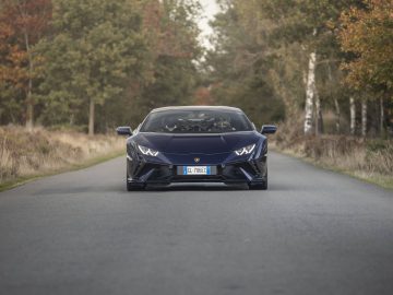 Een donkerblauwe Lamborghini-sportwagen staat centraal geparkeerd op een smalle, met bomen omzoomde weg met herfstbladeren op de achtergrond.