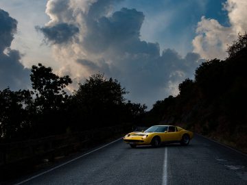 Een gele Lamborghini staat geparkeerd op een kronkelige bergweg onder een dramatische bewolkte hemel, die doet denken aan een scène uit een klassieke film. Bomen en vegetatie langs de weg.