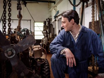 Een man in een blauwe jumpsuit zit geknield en inspecteert machines in een industriële werkplaats, net als een scène uit een film over het nauwgezette vakmanschap dat nodig is om een Lamborghini te maken.