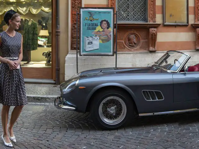 Een vrouw in een zwart-witte jurk met stippen staat naast een vintage grijze cabriolet op een geplaveide straat, wat de glamour oproept van een oude filmset met een gebouw en een poster op de achtergrond.