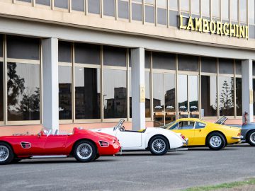 Vier vintage sportwagens, waaronder een rode, een witte, een gele en een grijze, staan geparkeerd voor een Lamborghini-dealer en zien eruit alsof ze regelrecht uit een klassieke filmset zijn gereden.