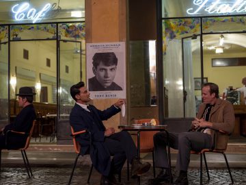 Twee mannen zitten aan een cafétafel voor een gebouw met de opschriften "Café" en "Italia", met op de achtergrond een zwart-wit poster van een jonge man genaamd Tony Benis aan de muur. Een andere man, die eruitziet alsof hij uit een oude film Lamborghini-advertentie is gestapt, zit alleen vlakbij.