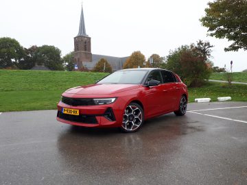 Een rode Opel Astra geparkeerd op een lege parkeerplaats op een natte dag, met een kerk en groen zichtbaar op de achtergrond. De auto heeft een gele kentekenplaat en opvallende lichtmetalen velgen, wat het strakke ontwerp benadrukt.