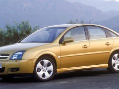 Een gouden Opel Vectra sedan staat geparkeerd op een open weg met groene struiken en bergen op de achtergrond.