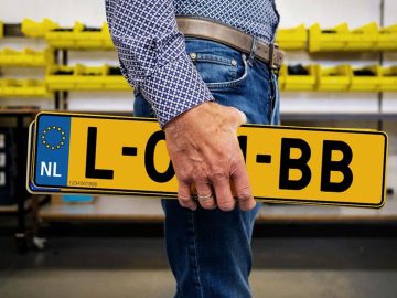 Een persoon in een geruit overhemd en spijkerbroek houdt een geel NL-kenteken vast met de code "L-001-BB" in een werkplaats vol gele opslagbakken. De kentekens zijn netjes geordend rond de werkruimte, wat bijdraagt aan de ordelijke omgeving.