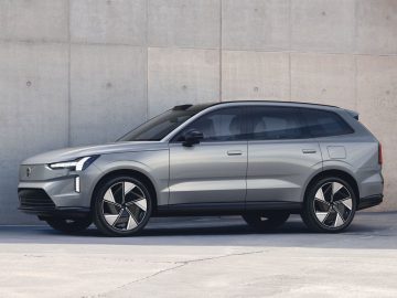 Een zilveren Volvo EX90 SUV staat geparkeerd voor een betonnen muur. Het voertuig beschikt over moderne designelementen, waaronder strakke lijnen en grote velgen.