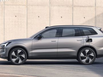 Een zilveren Volvo EX90 SUV staat geparkeerd naast een betonnen muur en laadt op bij een station. De deur van de oplaadpoort van het voertuig is open en de oplader is aangesloten.