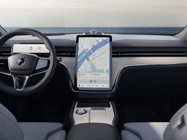 Binnenaanzicht van een Volvo EX90 met een strak stuur, een groot verticaal touchscreen met een kaart en een minimalistisch dashboardontwerp met een oceaanachtergrond zichtbaar door de voorruit.