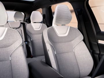 De afbeelding toont het interieur van een Volvo EX90 met drie rijen grijze stoffen stoelen met witte accenten. De stoelen zijn voorzien van hoofdsteunen en het moderne en strakke ontwerp weerspiegelt het verfijnde vakmanschap van de auto.