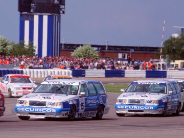 Een groep raceauto's, waaronder meerdere Volvo 850-breaks, navigeren een bocht op een racecircuit met een menigte op de achtergrond en een groot blauw-wit gebouw in de buurt.