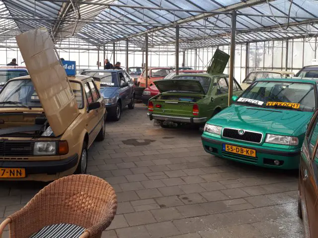 Een verscheidenheid aan auto's met open motorkap en kofferbak worden tentoongesteld in een grote kasachtige ruimte op Youngtimerworld. Een aantal auto's heeft gele kentekenplaten. Op de voorgrond is een rieten stoel zichtbaar.