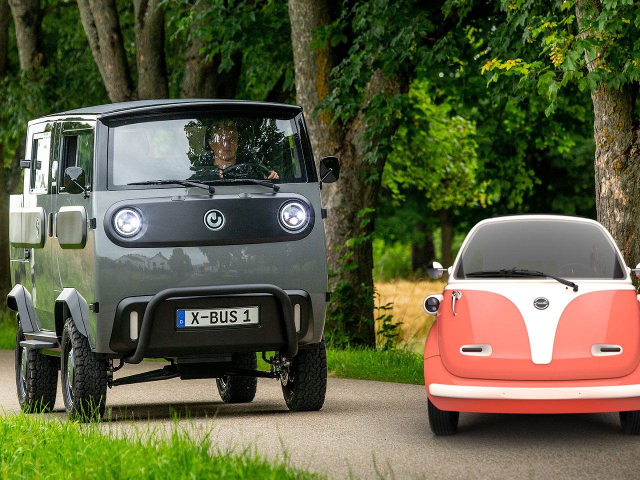 Twee kleine, uniek ontworpen elektrische voertuigen, een grijze XBUS en een rood-witte Microlino, staan geparkeerd op een landelijk pad omzoomd met bomen.