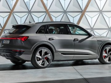 Een slanke grijze Audi Q8 e-tron SUV staat binnen geparkeerd tegen een muur met geometrische patronen. Het ontwerp wordt gekenmerkt door scherpe lijnen en stijlvolle lichtmetalen velgen.