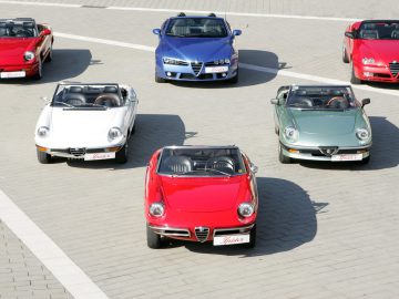 Op een verhard terrein staan in een halfronde opstelling meerdere cabriolets, waaronder een opvallende Alfa Romeo Spider – alles wat je moet weten – in diverse kleuren als rood, wit en groen.