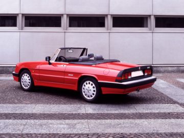 Een rode Alfa Romeo Spider converteerbare sportwagen staat geparkeerd op een geplaveide ondergrond voor de gevel van een modern gebouw.