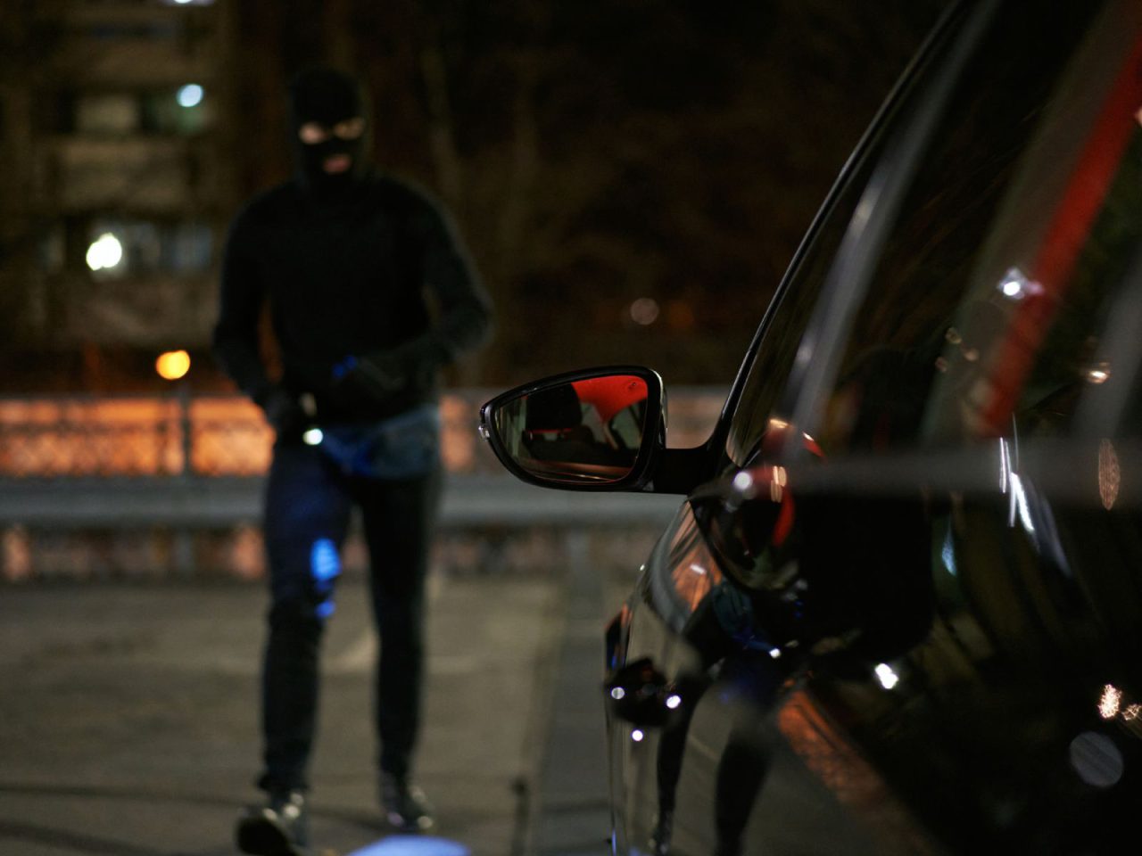 Een gemaskerd katalysatordief, gekleed in donkere kleding, nadert 's nachts een geparkeerde auto.