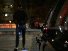 Een gemaskerd katalysatordief, gekleed in donkere kleding, nadert 's nachts een geparkeerde auto.