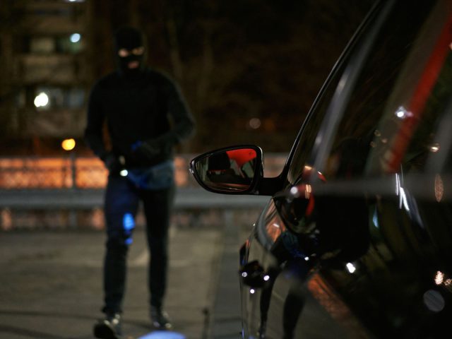 Een gemaskerd katalysatordief, gekleed in donkere kleding, nadert 's nachts een geparkeerde auto.