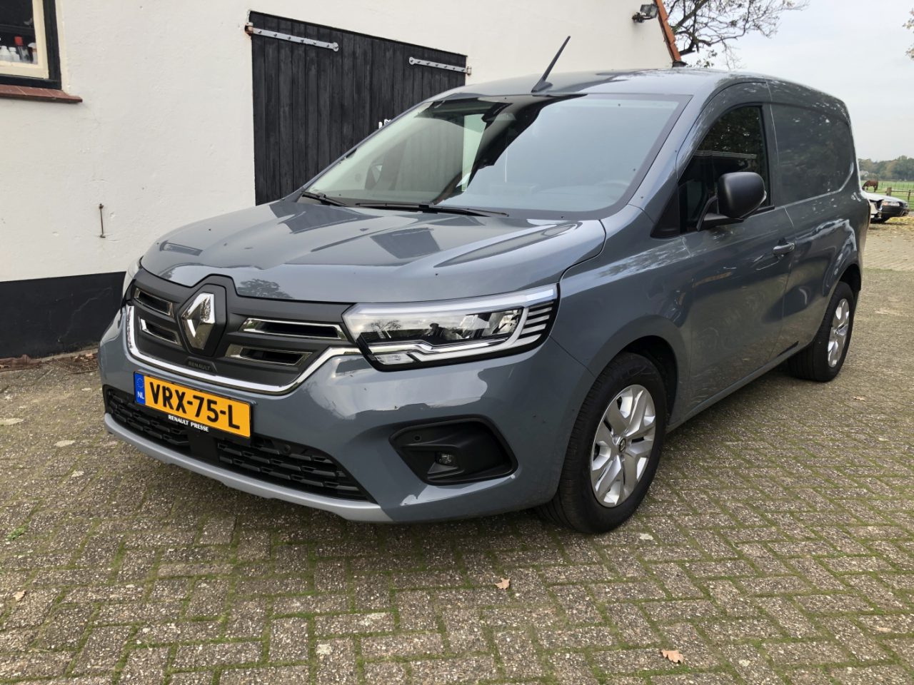 Een grijze Renault Kangoo-bestelwagen staat geparkeerd op een verharde ondergrond voor een wit-zwart gebouw. Het voertuig heeft een geel kenteken met kenteken VRX-75-L.